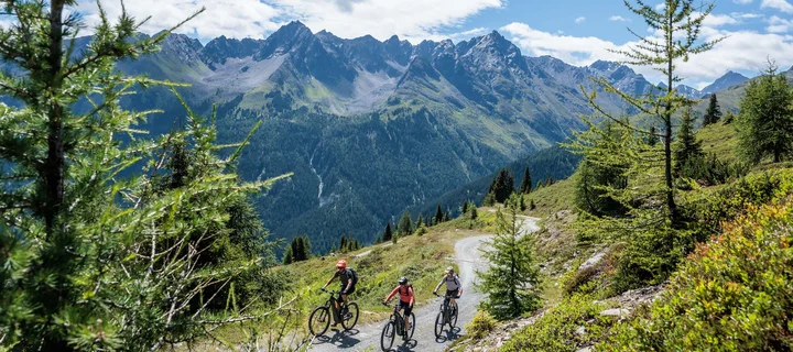 Ferienglück und Alpenland | Ihre stilvollen Hotels in Ischgl Drei Mountainbiker fahren auf einem Bergweg vor einer alpinen Bergkulisse