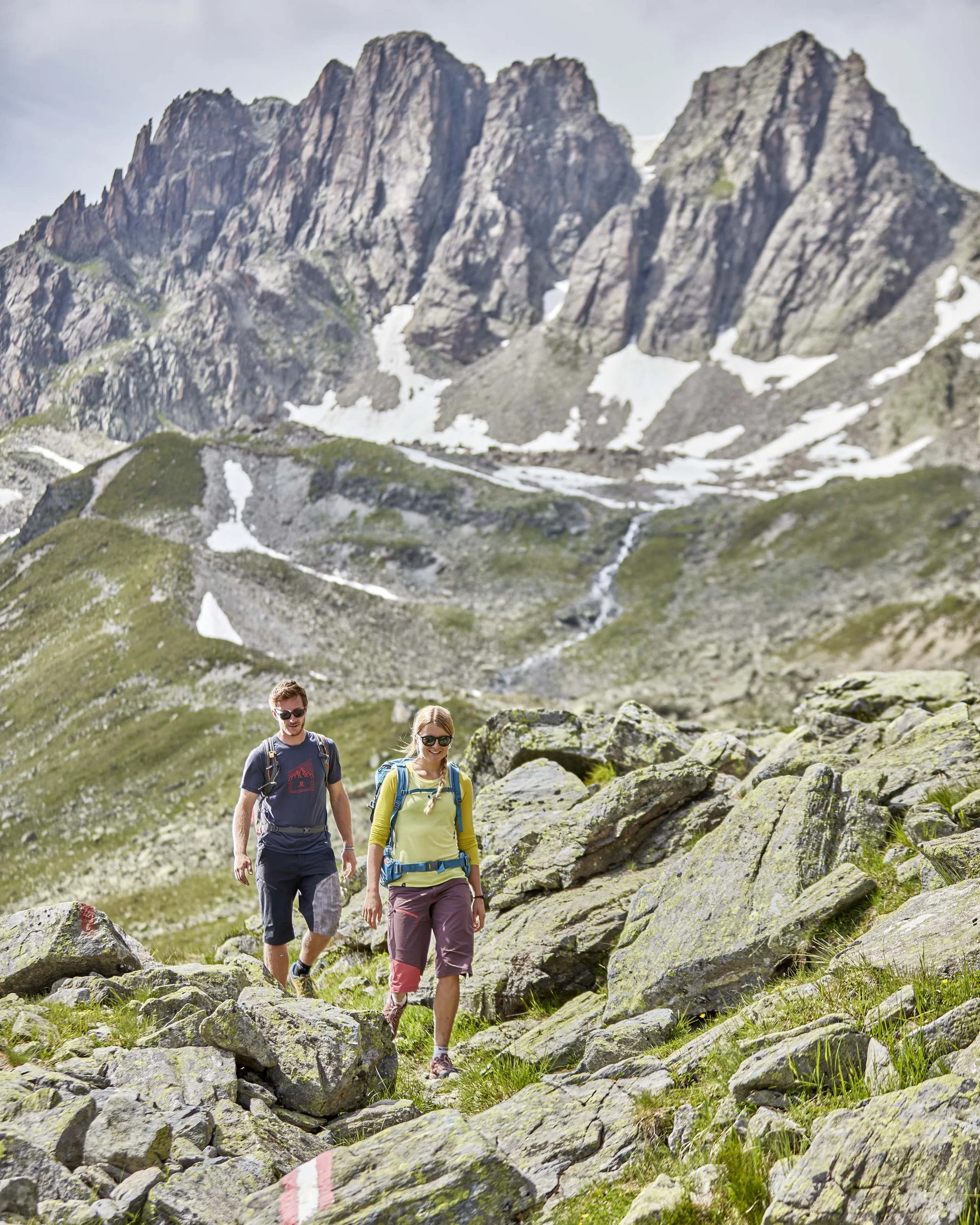 Ferienglück und Alpenland | Ihre stilvollen Hotels in Ischgl Zwei Wanderer auf felsigem Bergpfad vor schroffen Gipfeln mit Schneefeldern